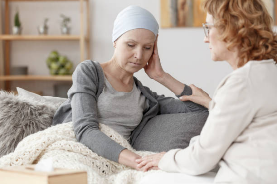 Woman with cancer wearing a head wrap sits beside her friend on the couch holding her hand.