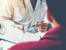 A doctor in a white lab coat holding a clipboard and a pen up to a patient.